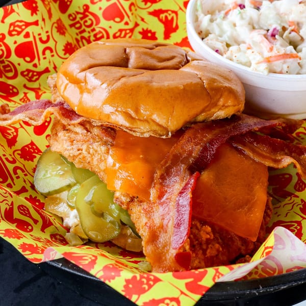Close-up of a fried chicken sandwich with cheese, bacon, and pickles, served with coleslaw.