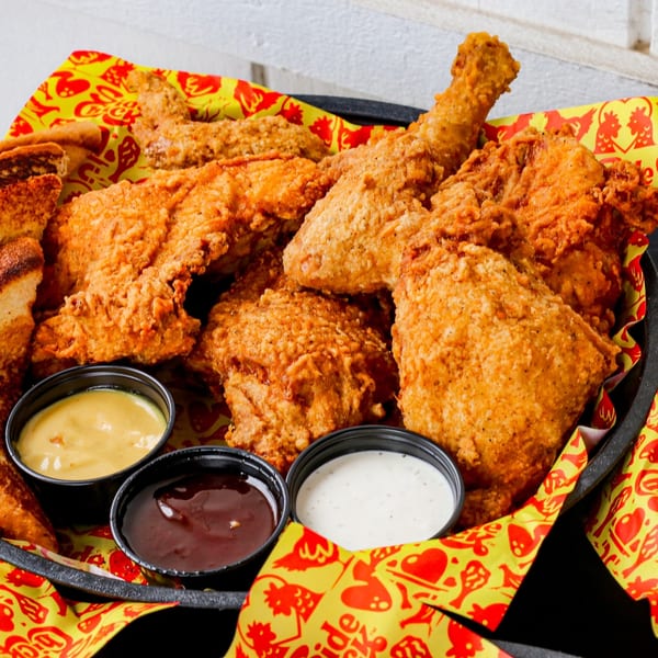 Crispy fried chicken pieces, toast, and three dipping sauces in a black basket lined with yellow paper.